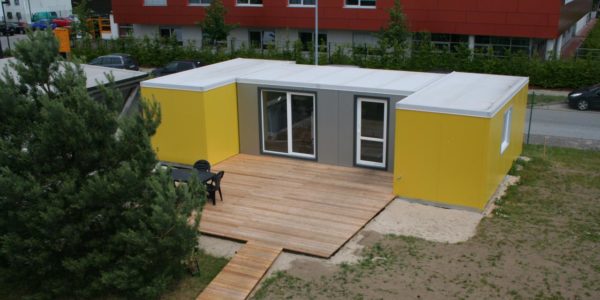 Ein kleines Wohngebäude aus Stahlsandwich-Elementen in grau und gelb mit großer Holzterasse und großen Fenstern