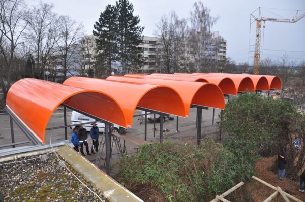 Leuchtend orange, halbrunde Dachelemente über einem großen Fahrradständer in einer urbanen Umgebung